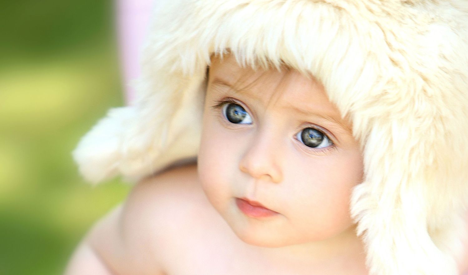 Adorable baby with striking blue eyes wearing a fluffy white hat in a soft focus outdoor portrait.
