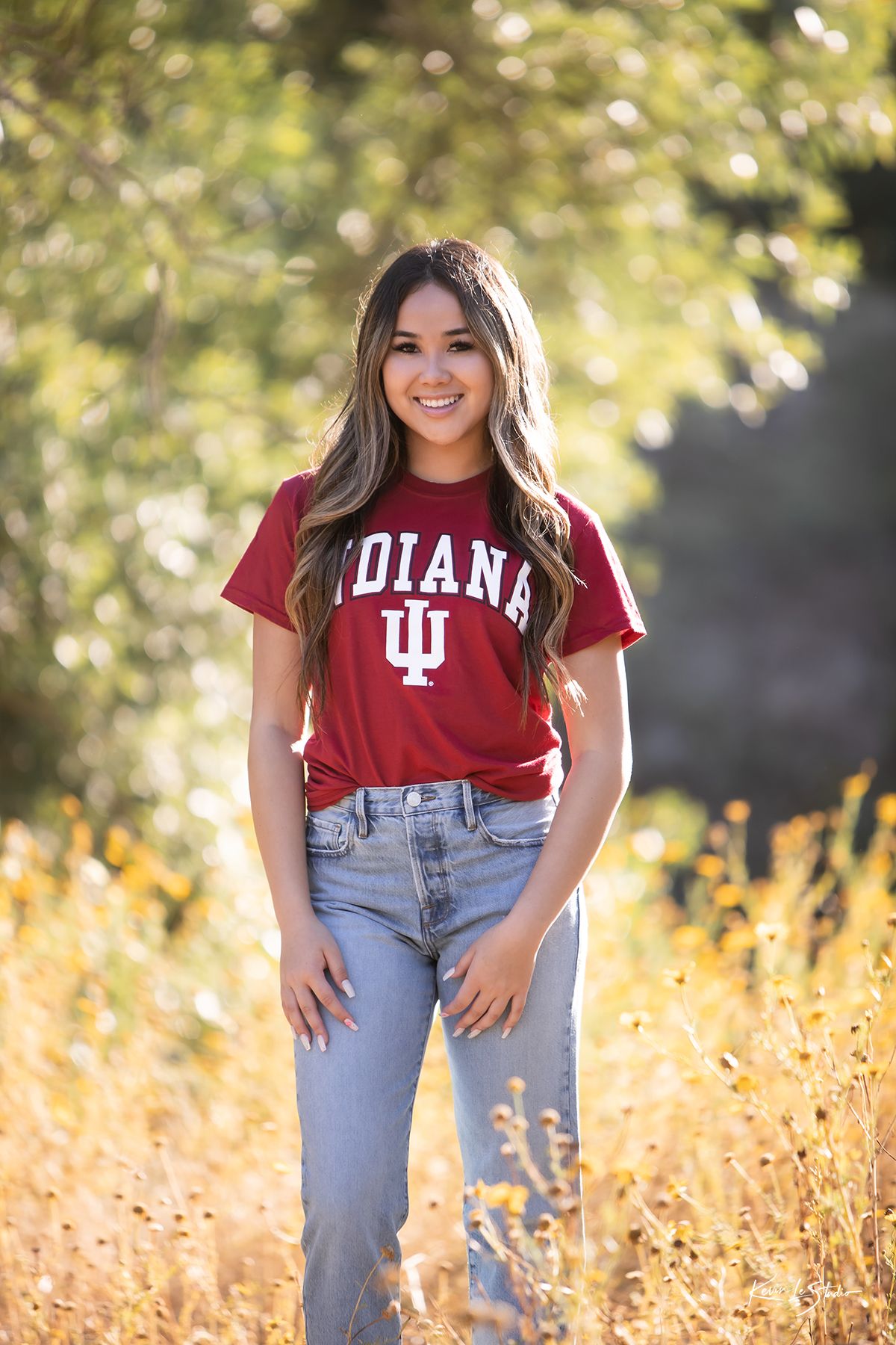 Cheerful young woman in Indiana University red t-shirt and jeans poses in golden meadow for senior portraits.