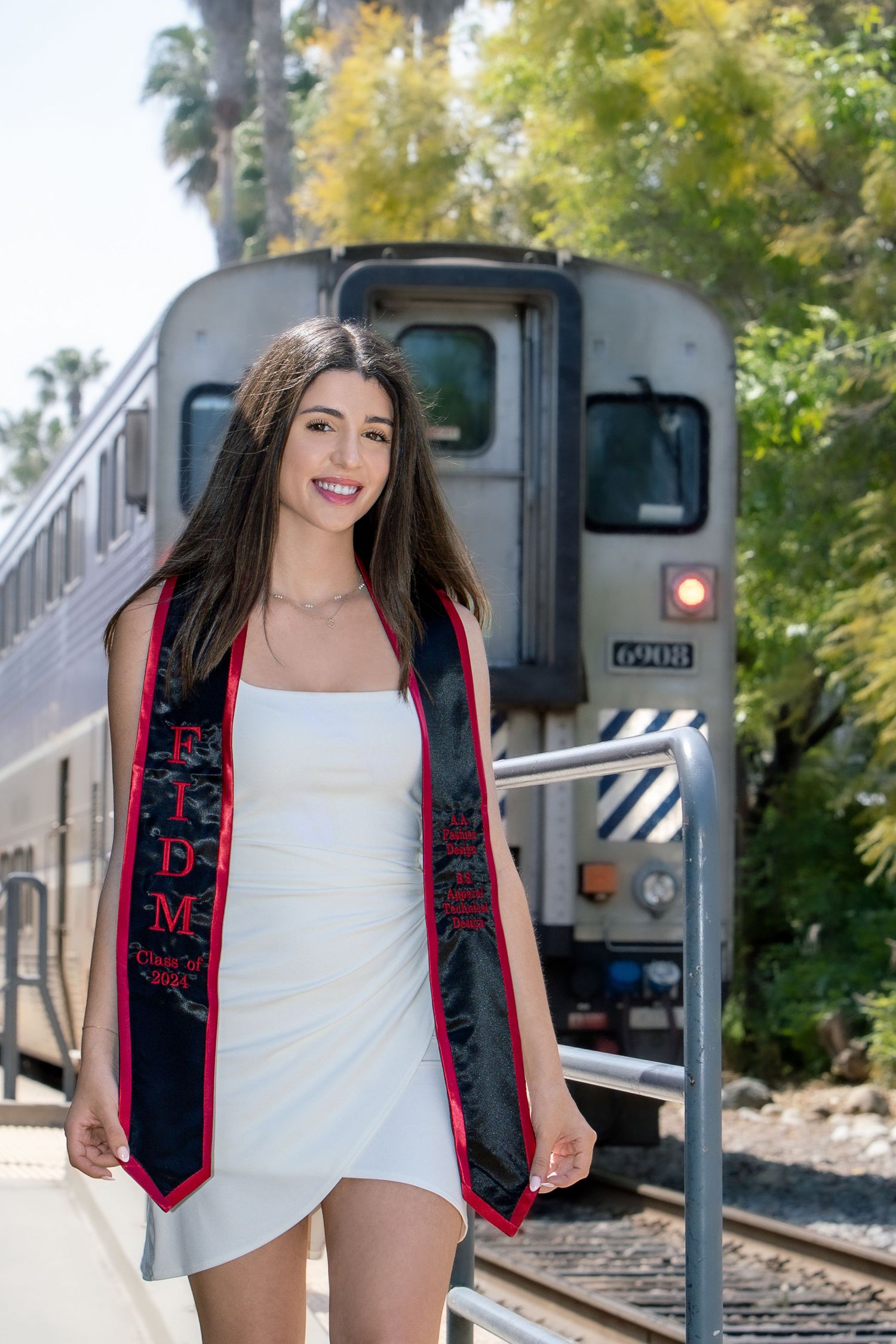 Female FIDM graduate in white dress with graduation stole poses confidently on train platform with locomotive behind.