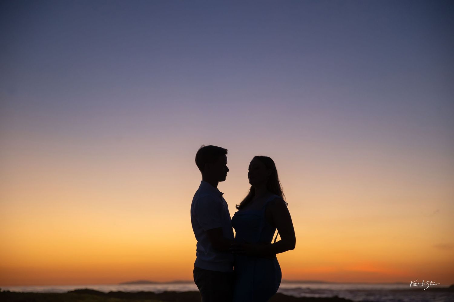 Romantic silhouette of a couple facing each other against a vibrant orange and blue sunset sky by the ocean.
