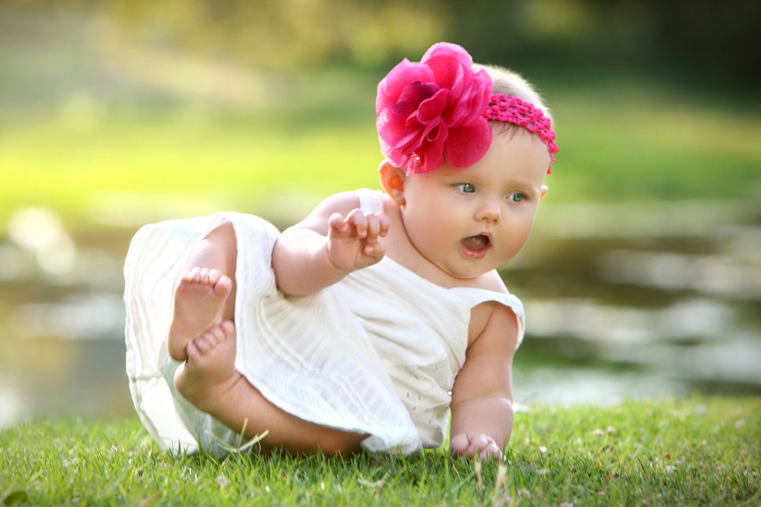 Adorable baby girl in white dress with pink flower headband crawling on green grass outdoors.