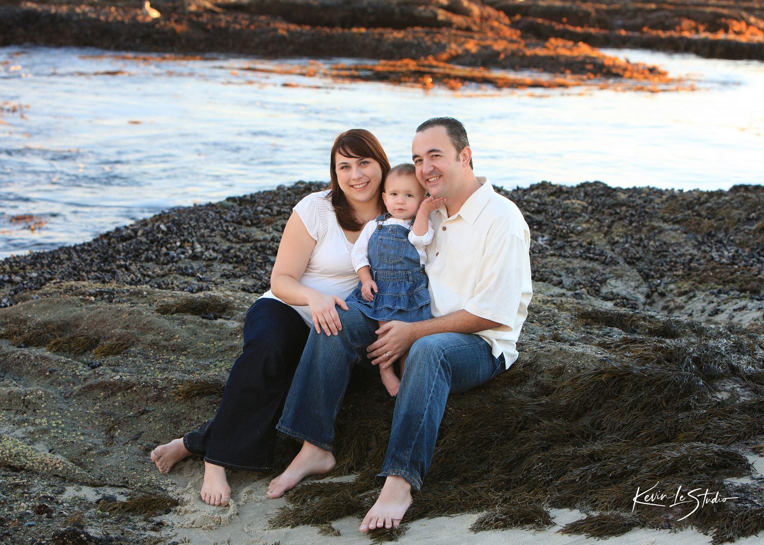 Family of three smiling together on rocky beach at sunset, parents sitting with toddler between them.