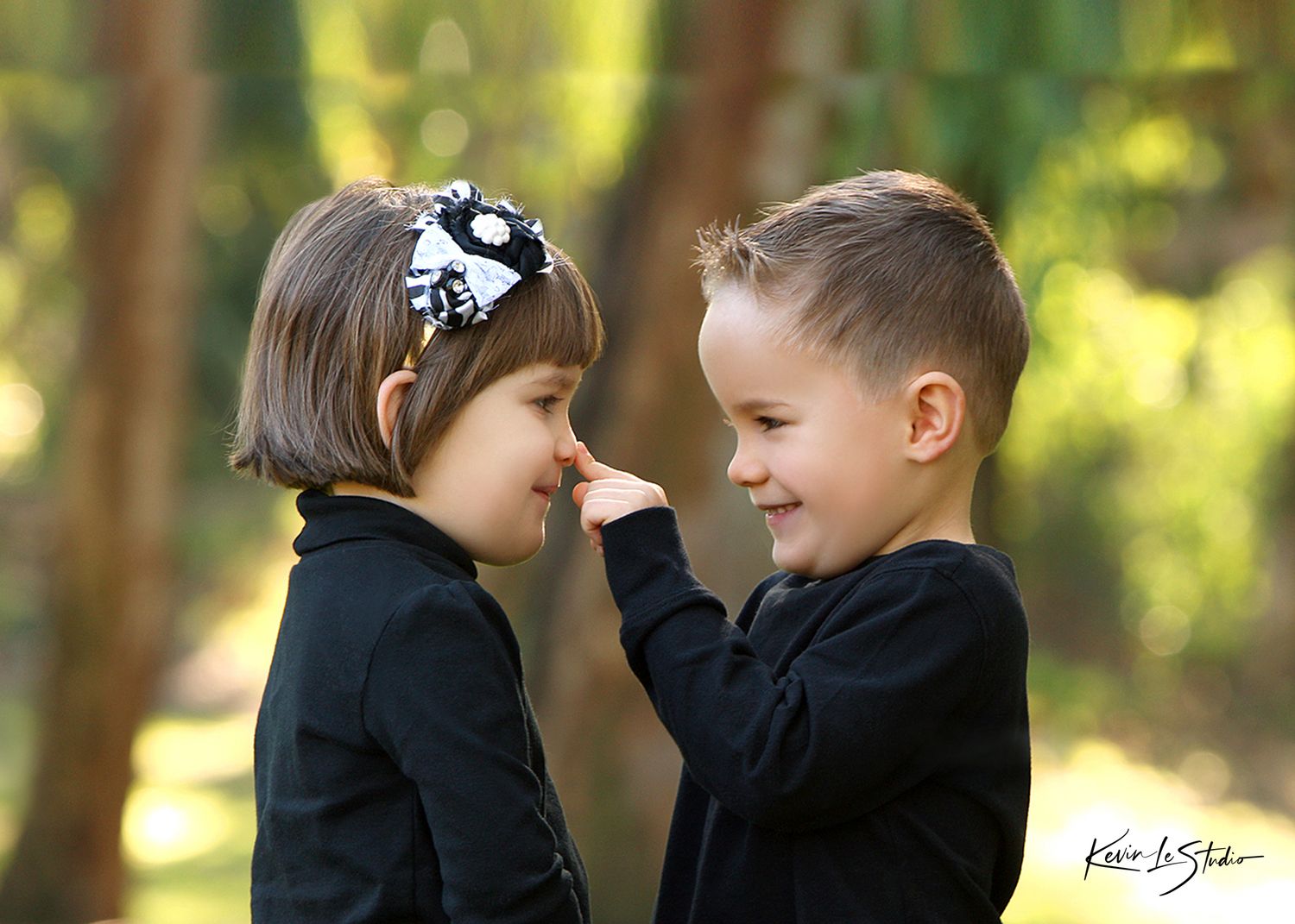 Two young children in black outfits share a sweet moment outdoors, with a boy gently touching a girl's nose.