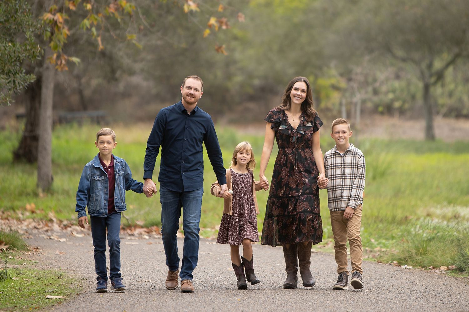 Family of five walking hand-in-hand on a park path, smiling outdoors in autumn greenery.