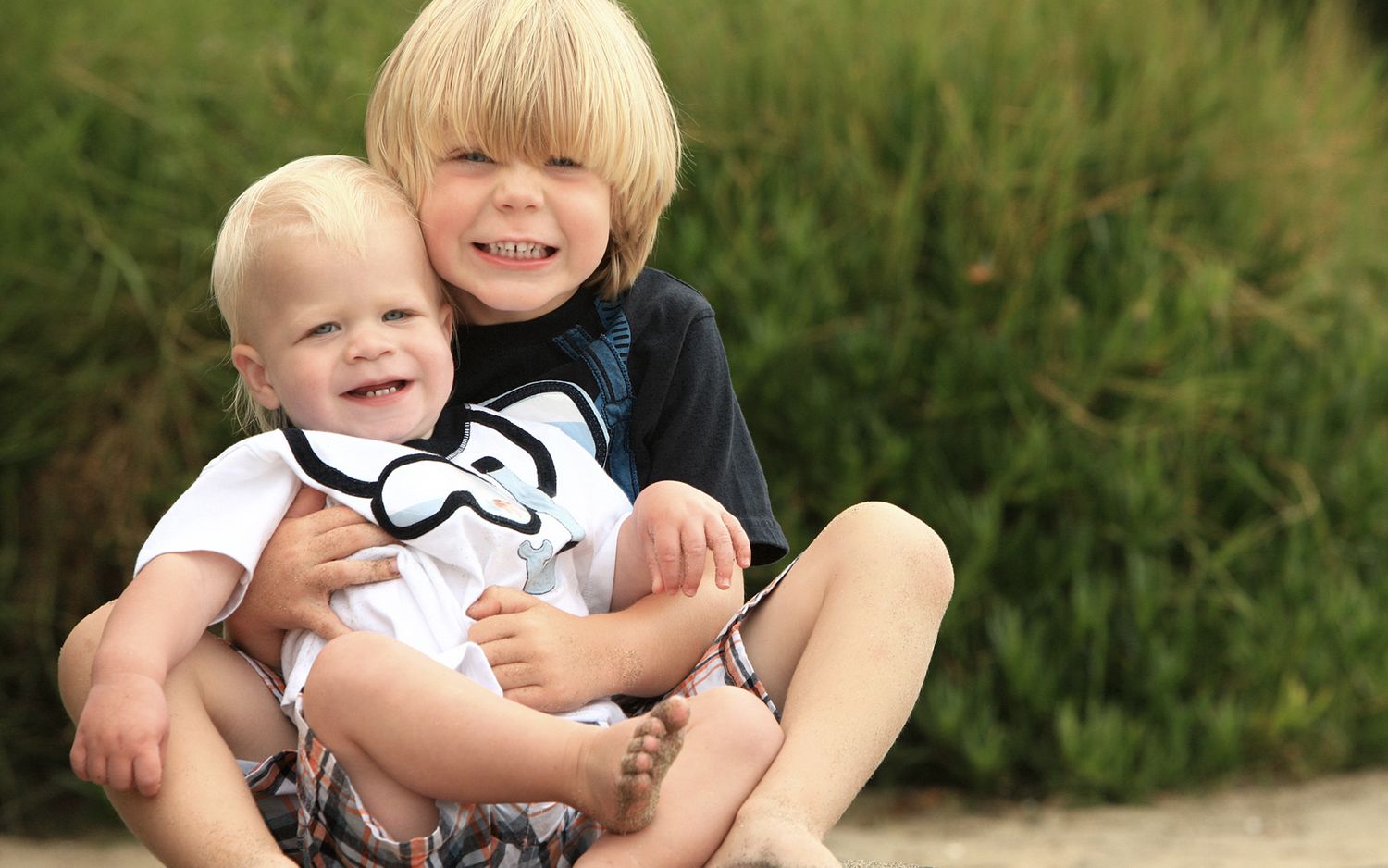 Two blonde boys sitting outdoors, older child hugging baby holding mustache prop, smiling happily on sandy ground.