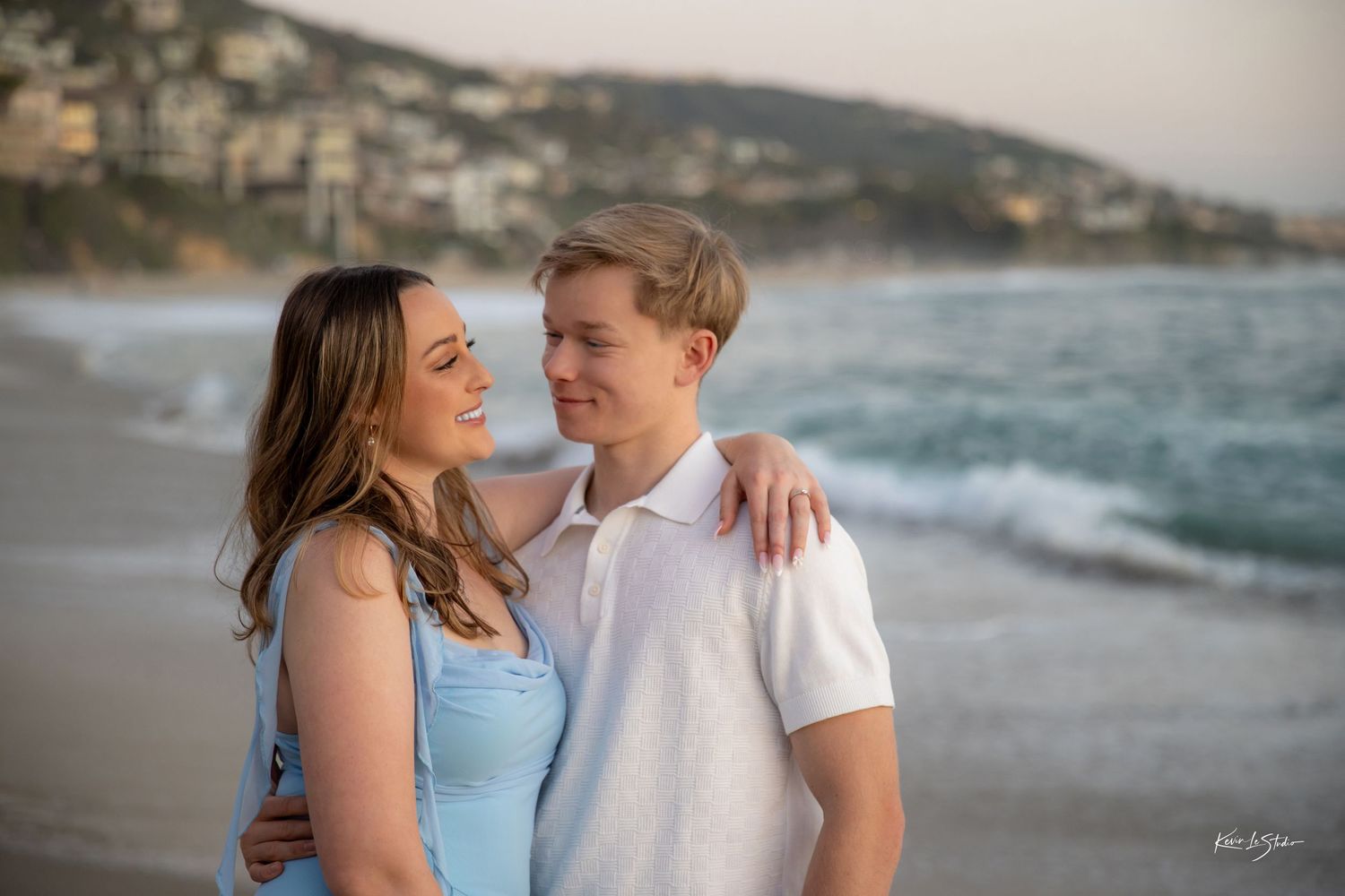 Smiling couple embracing on beach with coastal town in background at sunset.