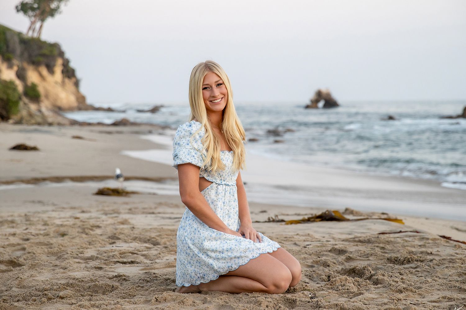 Young blonde woman in floral dress sitting on sandy beach with rocky cliffs in background.
