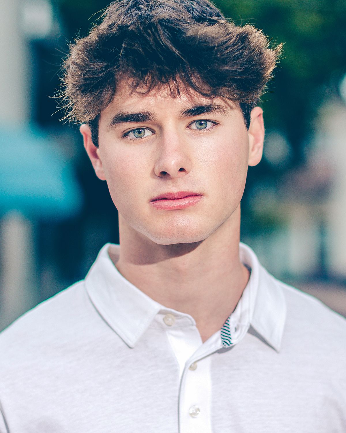 Young man with blue eyes in white polo shirt posing outdoors with blurred background.