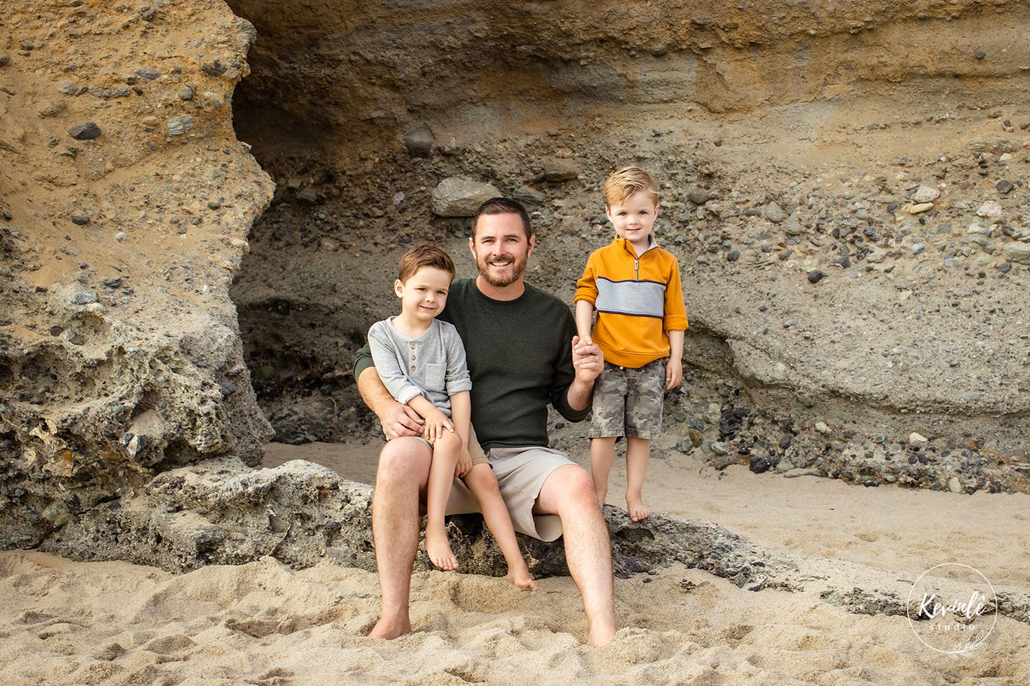 A man and two young children smile together in a sandy rock cave at the beach.
