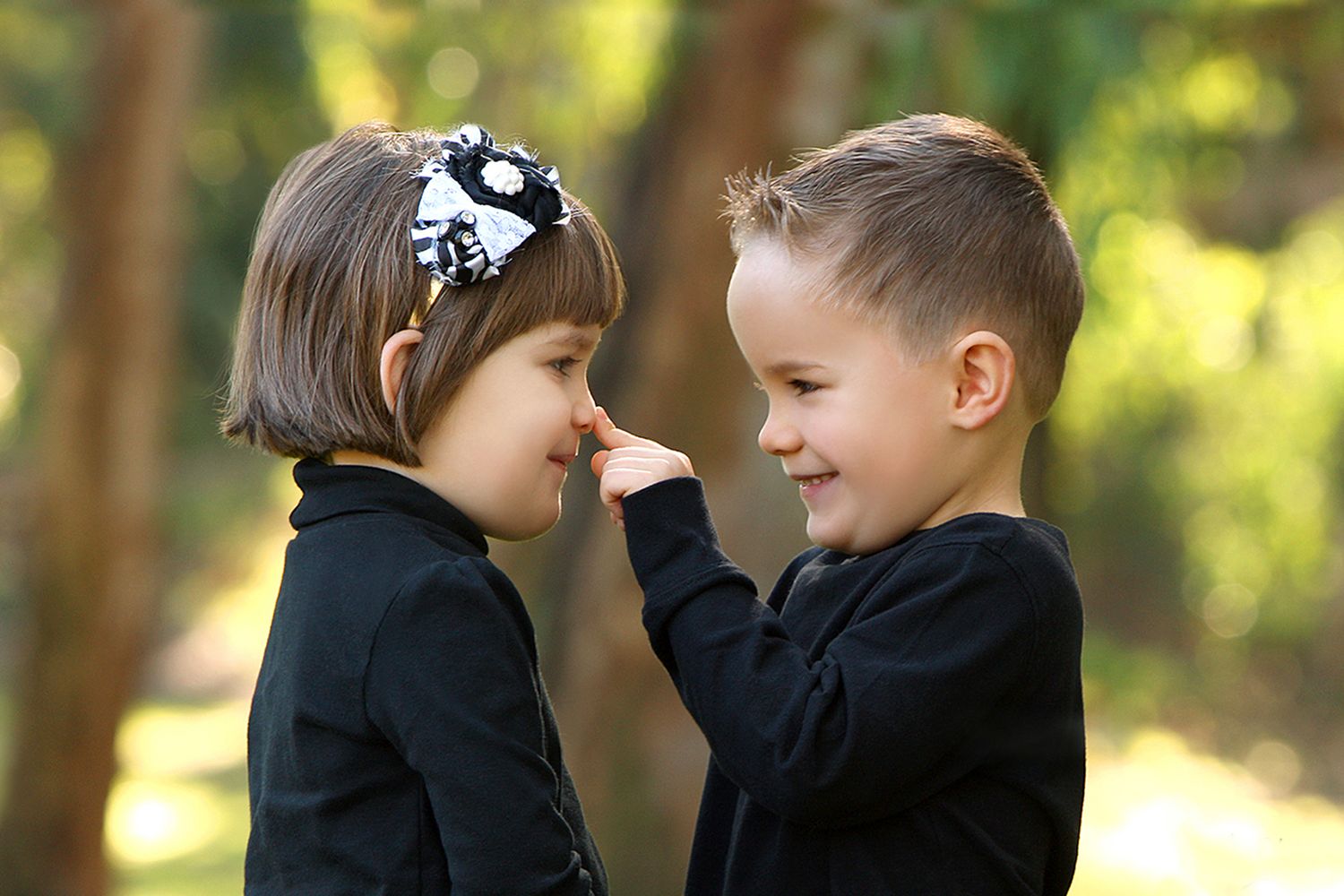 Two young children in matching black outfits share a playful moment outdoors, with a boy gently touching a girl's nose.