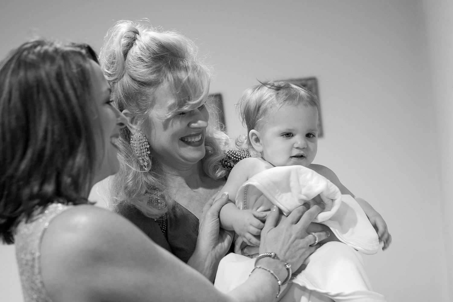 Black and white photo of a smiling woman holding a baby in a white outfit, with another woman nearby.