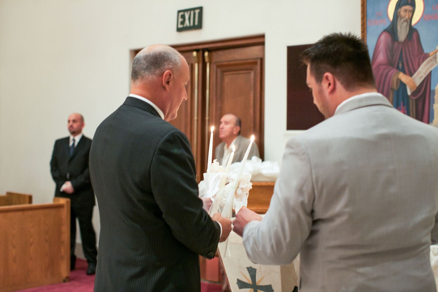 Two men exchanging items during a church ceremony with an exit sign and religious icon visible in the background.