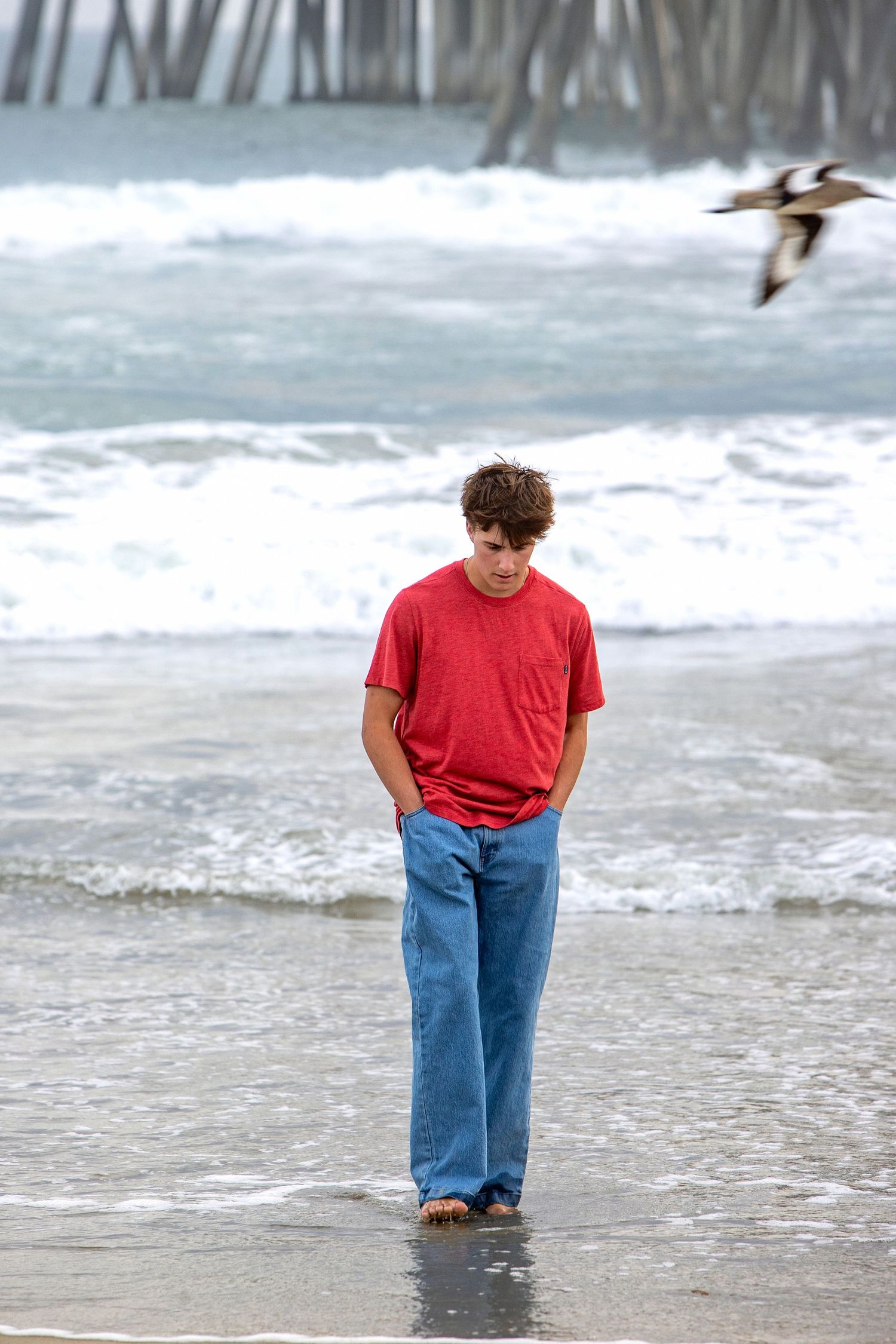 A teenage boy in a red t-shirt and jeans walks barefoot on a beach near a pier with waves and a bird in flight.