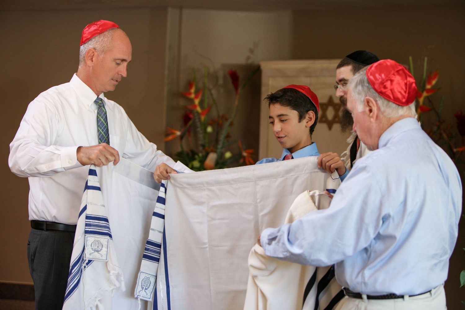 Men in red yarmulkes and tallits participate in a Jewish Bar Mitzvah ceremony indoors.