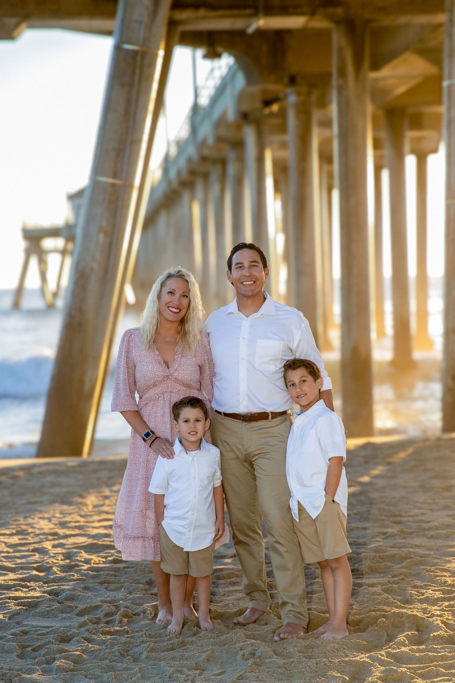 Family of four posing on sandy beach under wooden pier at golden hour, parents and two boys in coordinating outfits.