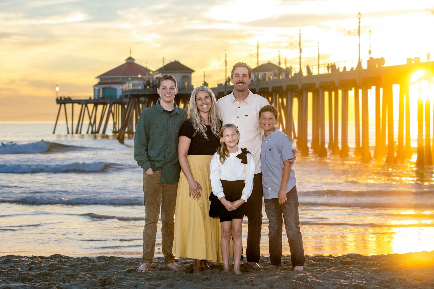 Family of five posing on beach at golden sunset near pier, parents with three children smiling.