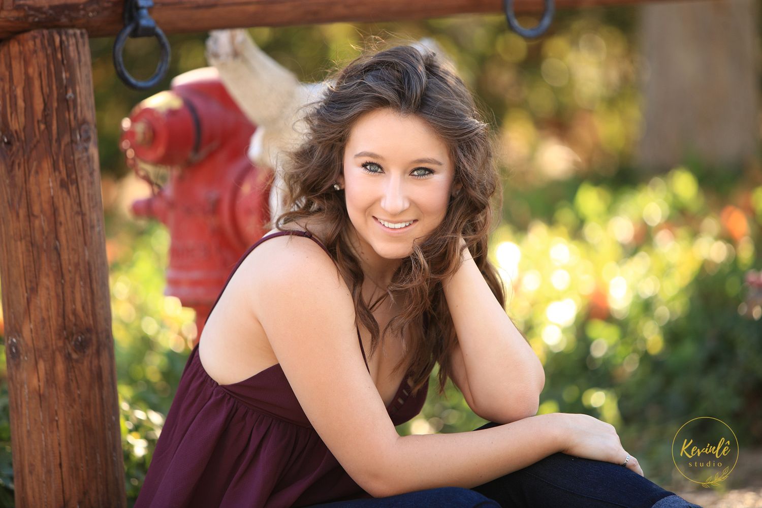 Young woman with curly hair in maroon top smiling outdoors near red fire hydrant in professional senior portrait.