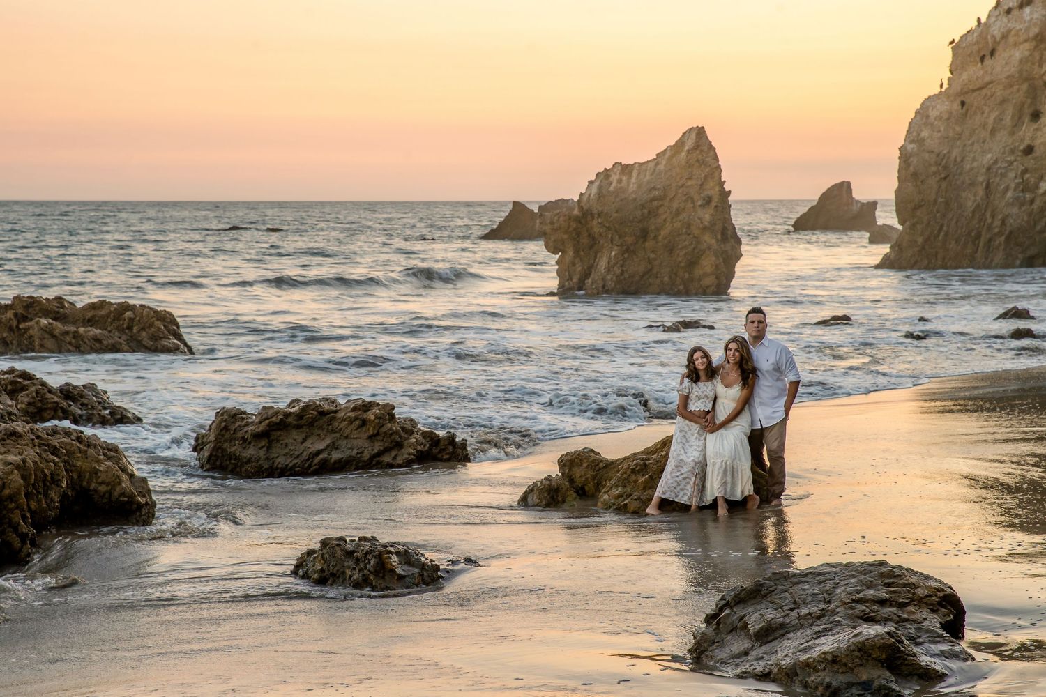 Family of three poses on rocky beach at sunset with golden pink sky and ocean waves at El Matador Beach.