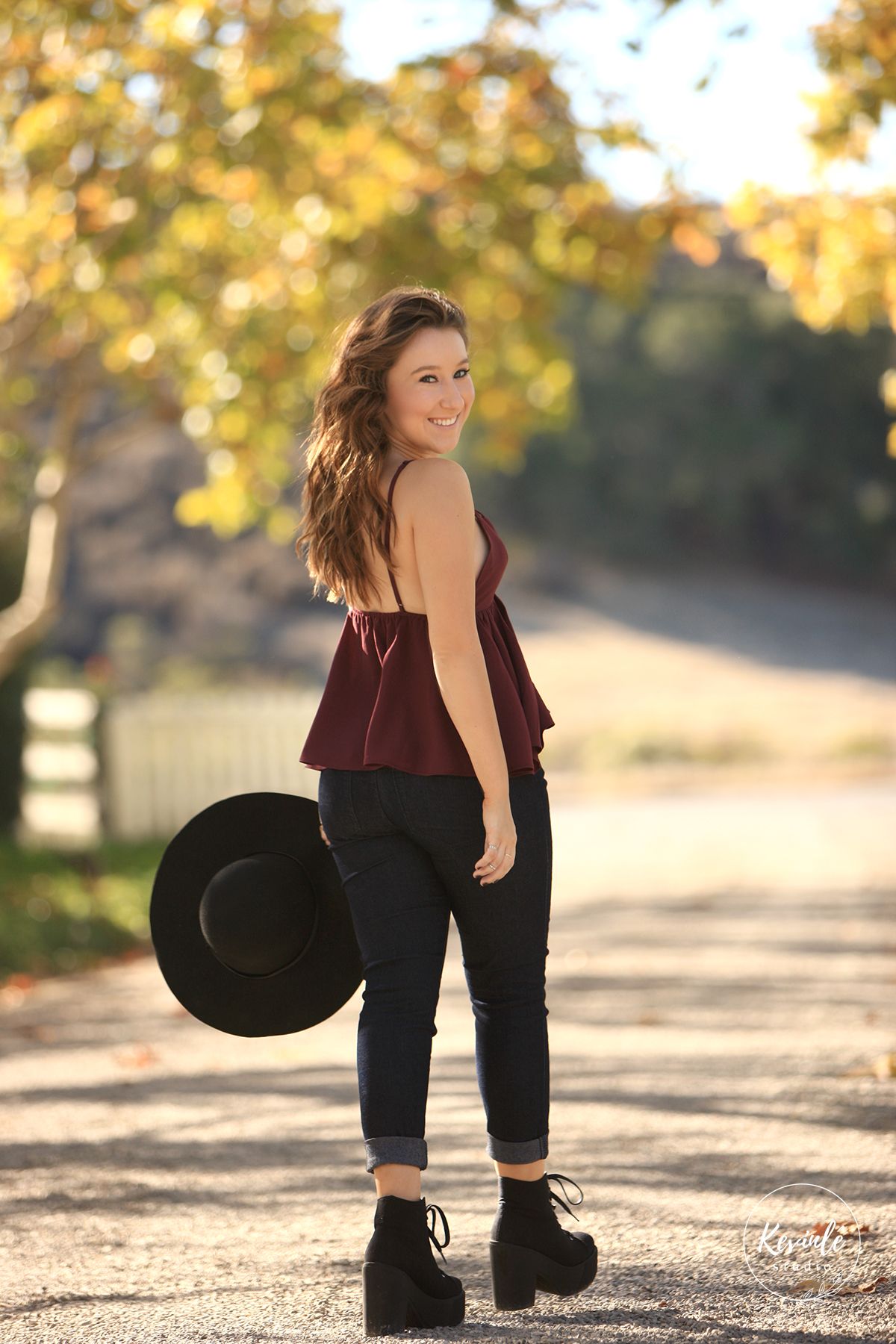 Young woman in burgundy top and black jeans holds a hat on a sunlit country road in autumn.