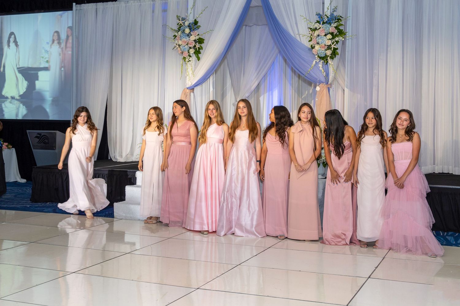 Young girls in pink and white formal gowns stand on a dance floor during an elegant pageant or quinceañera ceremony.