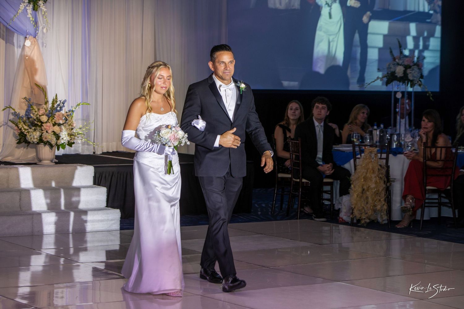 Bride in white gown and groom in black tuxedo make grand entrance at elegant wedding reception ballroom.