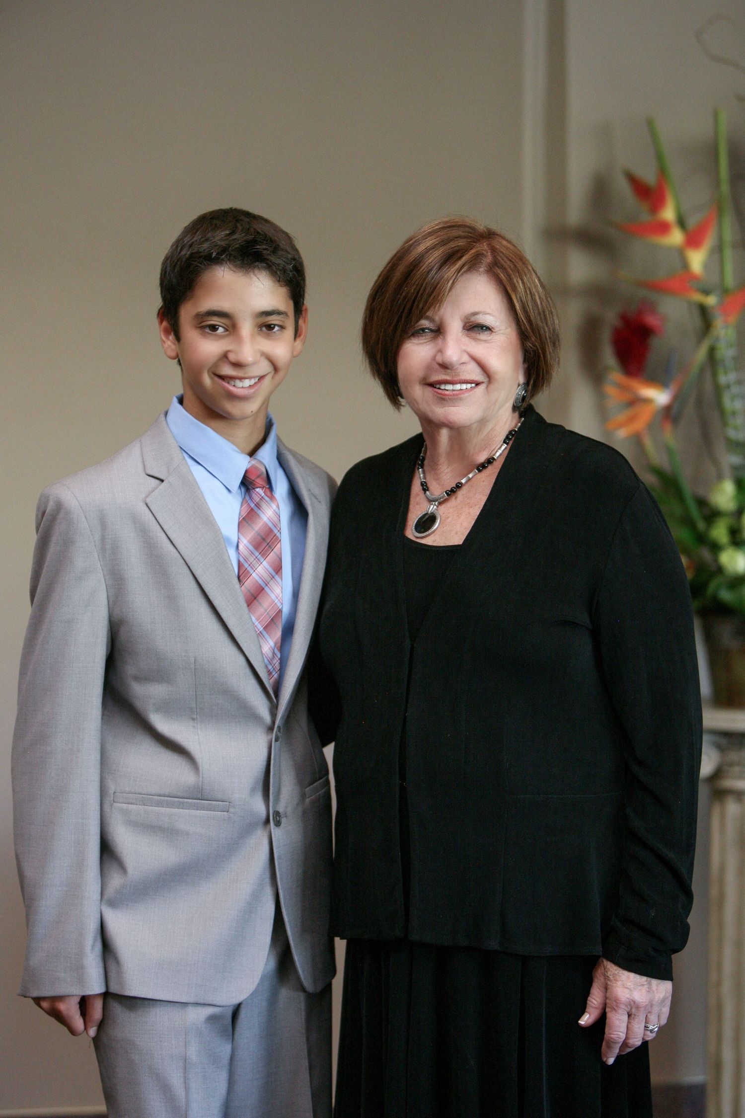 A young boy in a gray suit and tie poses with an older woman in black at a formal event.