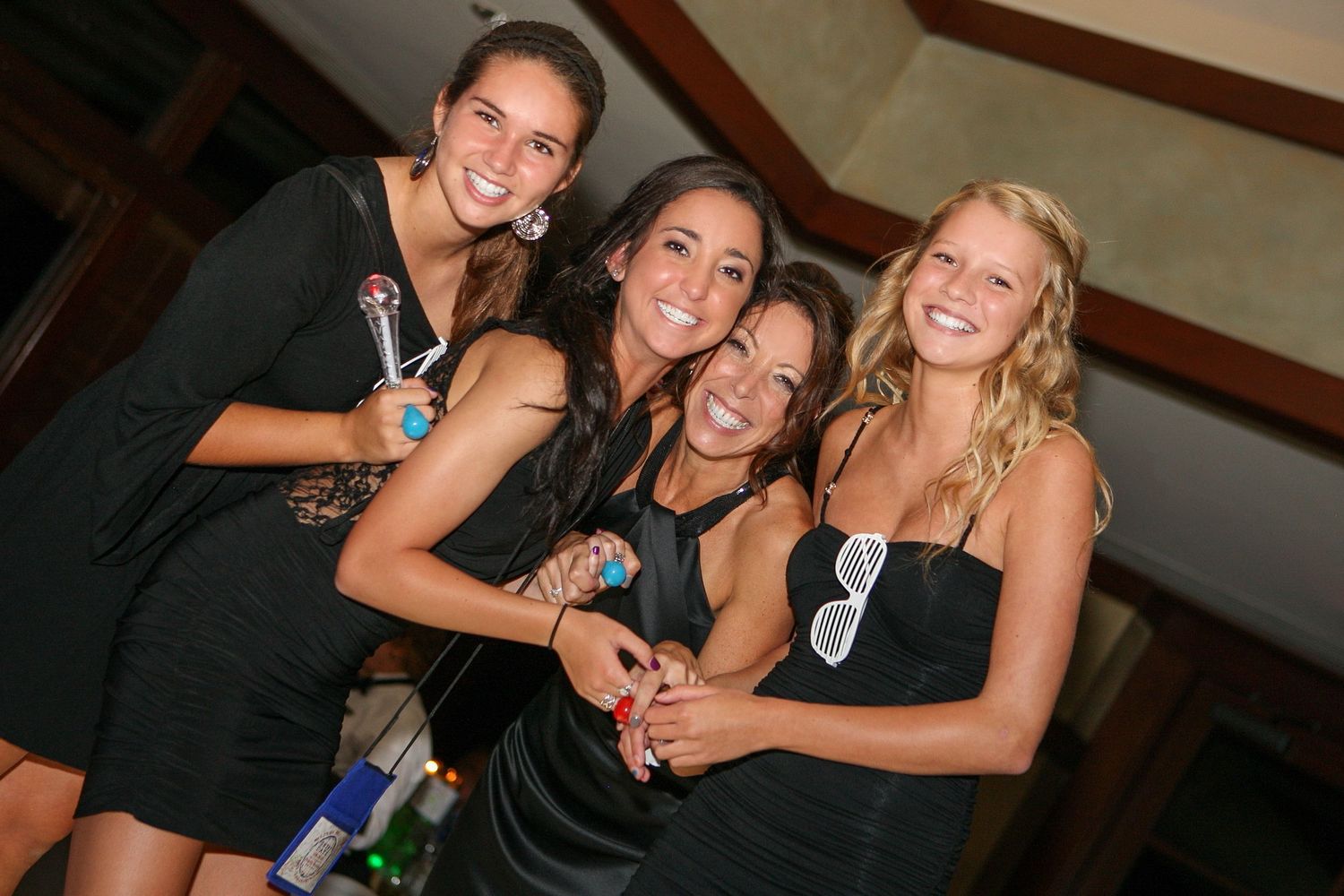 Four smiling young women in black dresses posing together at a social event or party.
