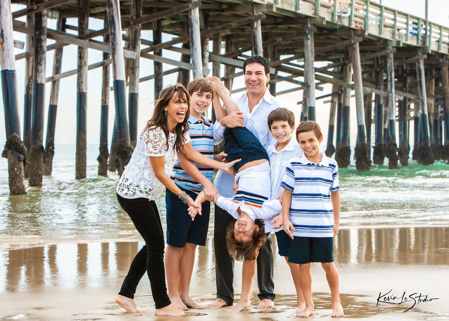 Family of six posing and laughing on sandy beach near wooden pier, with one child doing a handstand.