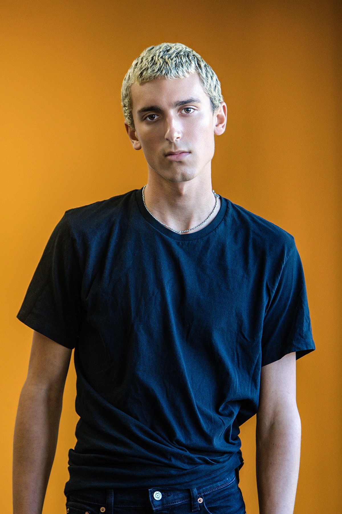 Young man with bleached blonde hair in black t-shirt poses against vibrant orange background.