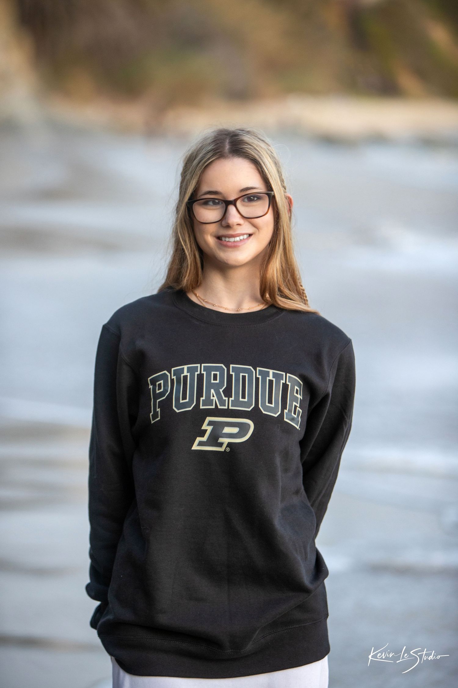 Smiling blonde teen in Purdue sweatshirt and glasses posing at beach.
