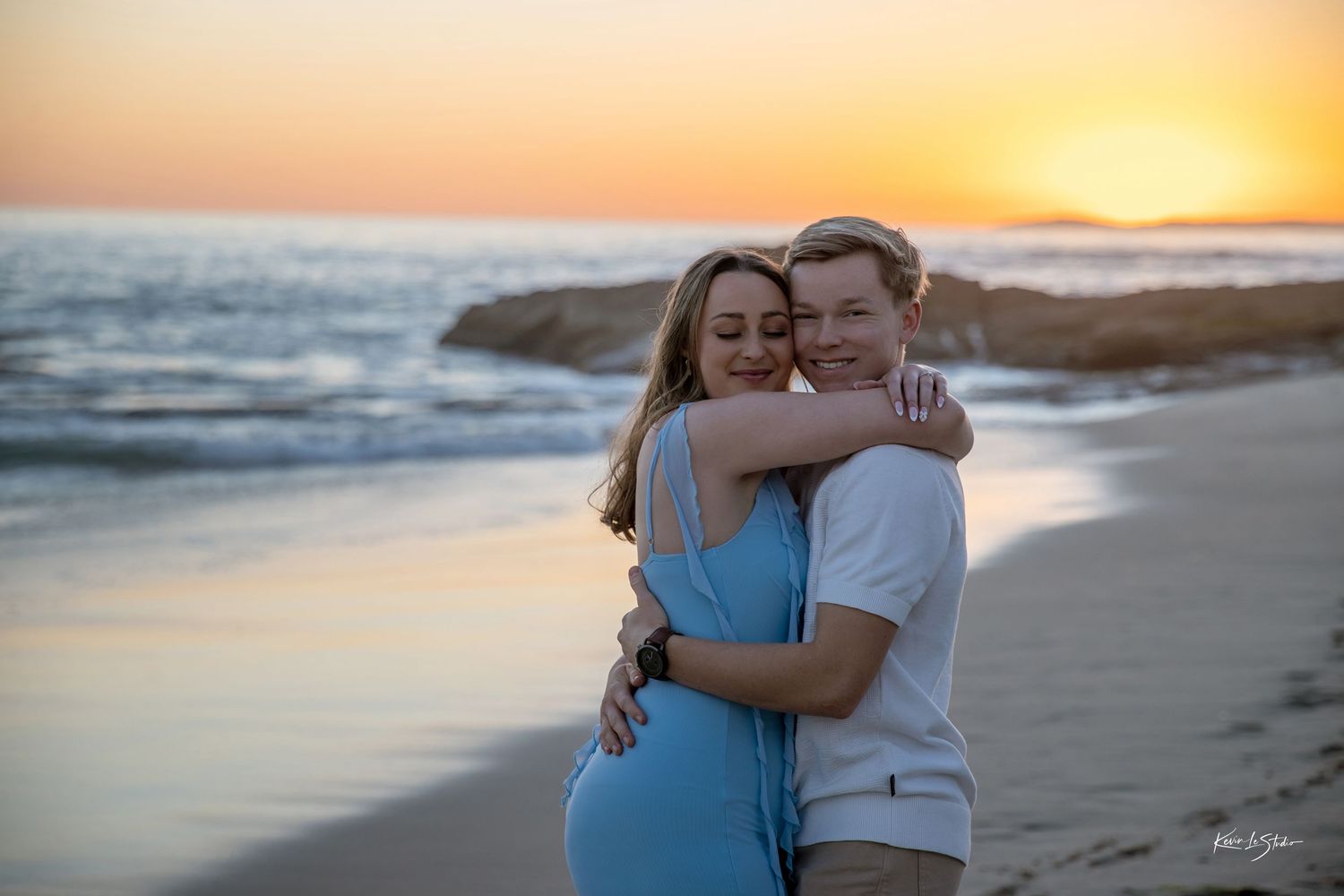 Couple embracing on beach at golden sunset, woman in blue dress hugging man in white shirt.