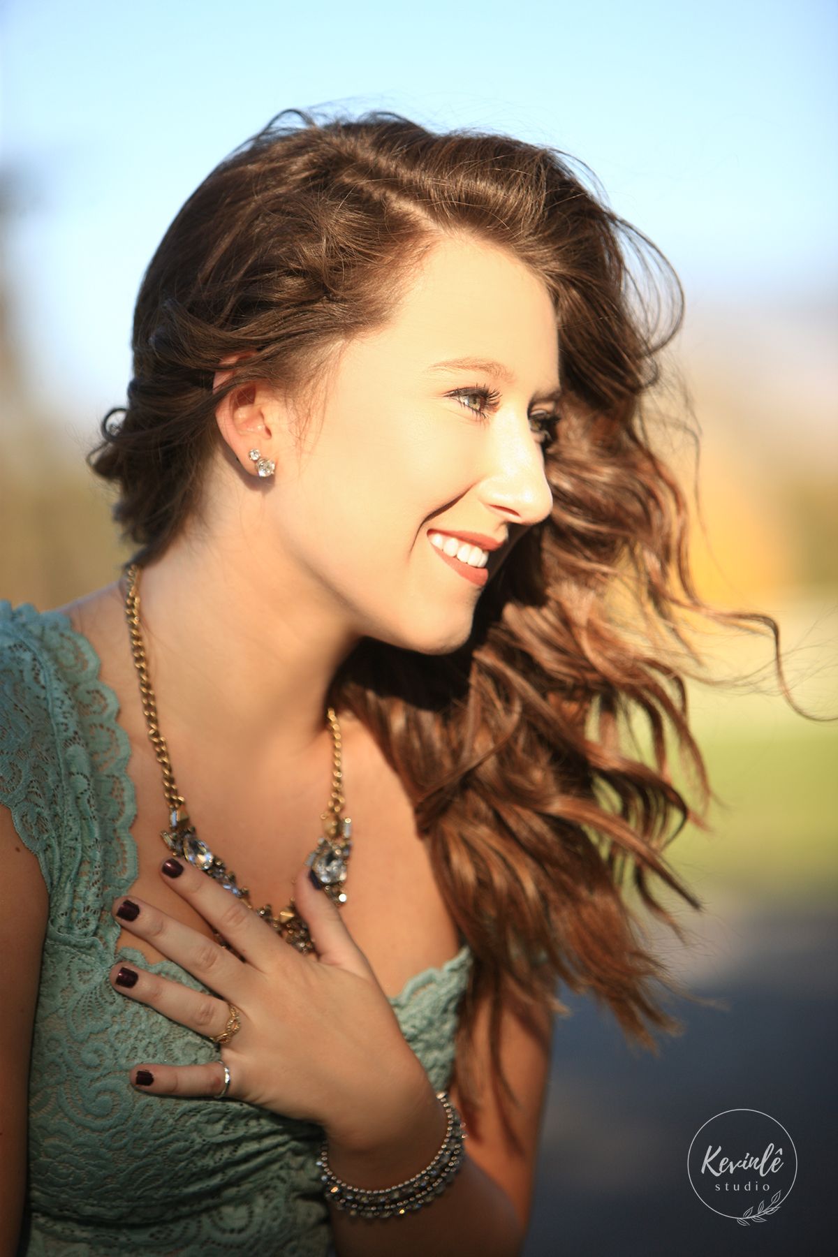 Young woman with curly hair smiling in warm sunlight wearing teal lace dress and jewelry.