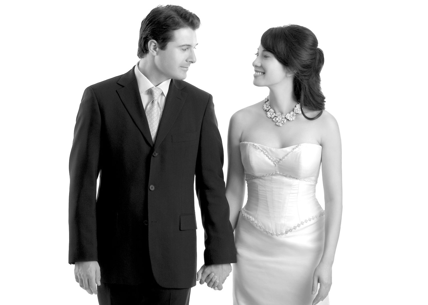 A couple in formal wedding attire, man in black suit and woman in white strapless gown, smiling at each other.