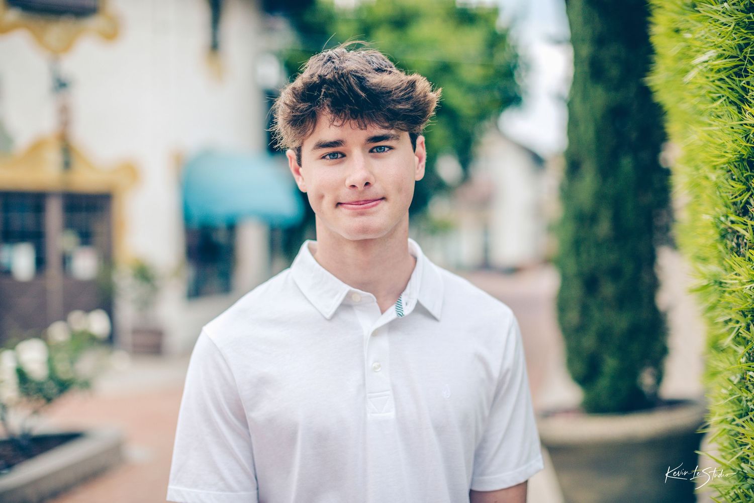 Smiling young man in white polo shirt posing outdoors near tall cypress trees in a village square.