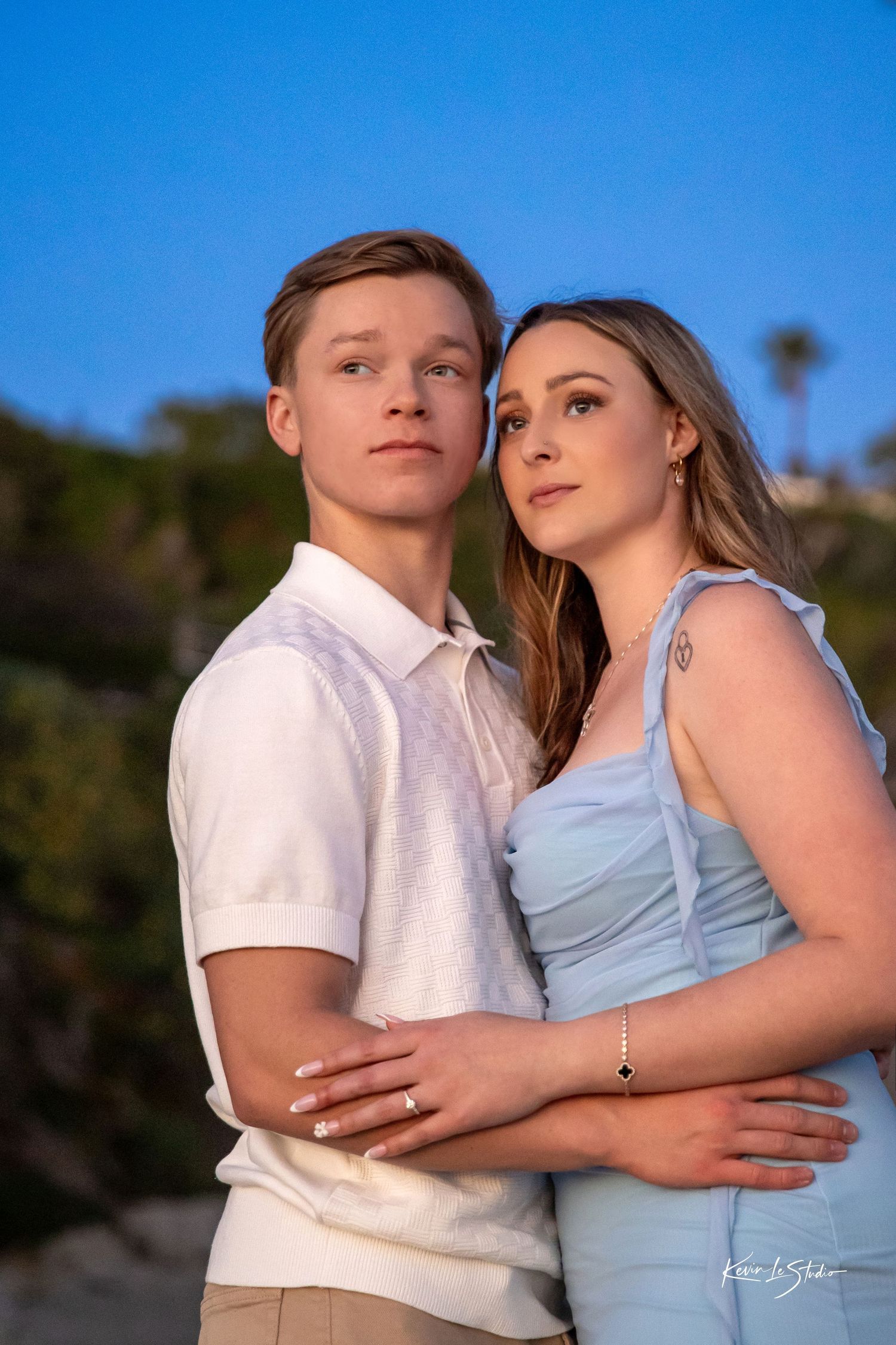 Young engaged couple posing outdoors at golden hour, woman in blue dress embracing man in white polo shirt.