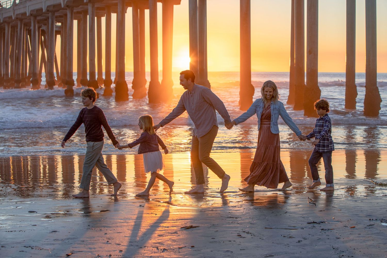 Family of five holds hands walking on beach at golden sunset near a pier silhouette.
