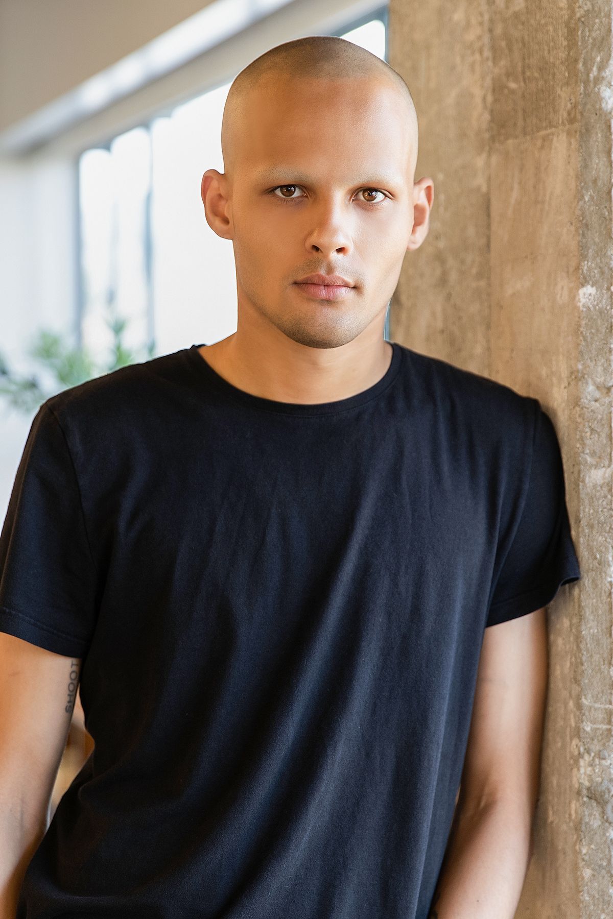 Bald young man wearing a black t-shirt, leaning against a concrete column with a serious expression.