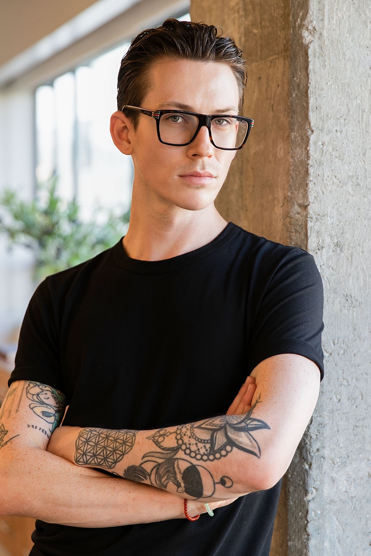 Young man with slicked-back hair, black-rimmed glasses, and tattoos wearing a black t-shirt with arms crossed.