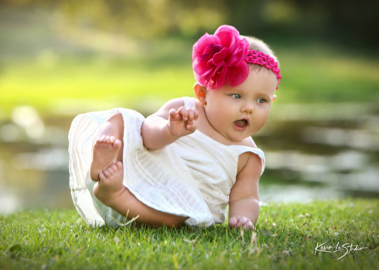Adorable baby girl in white dress and pink flower headband crawling on green grass outdoors.