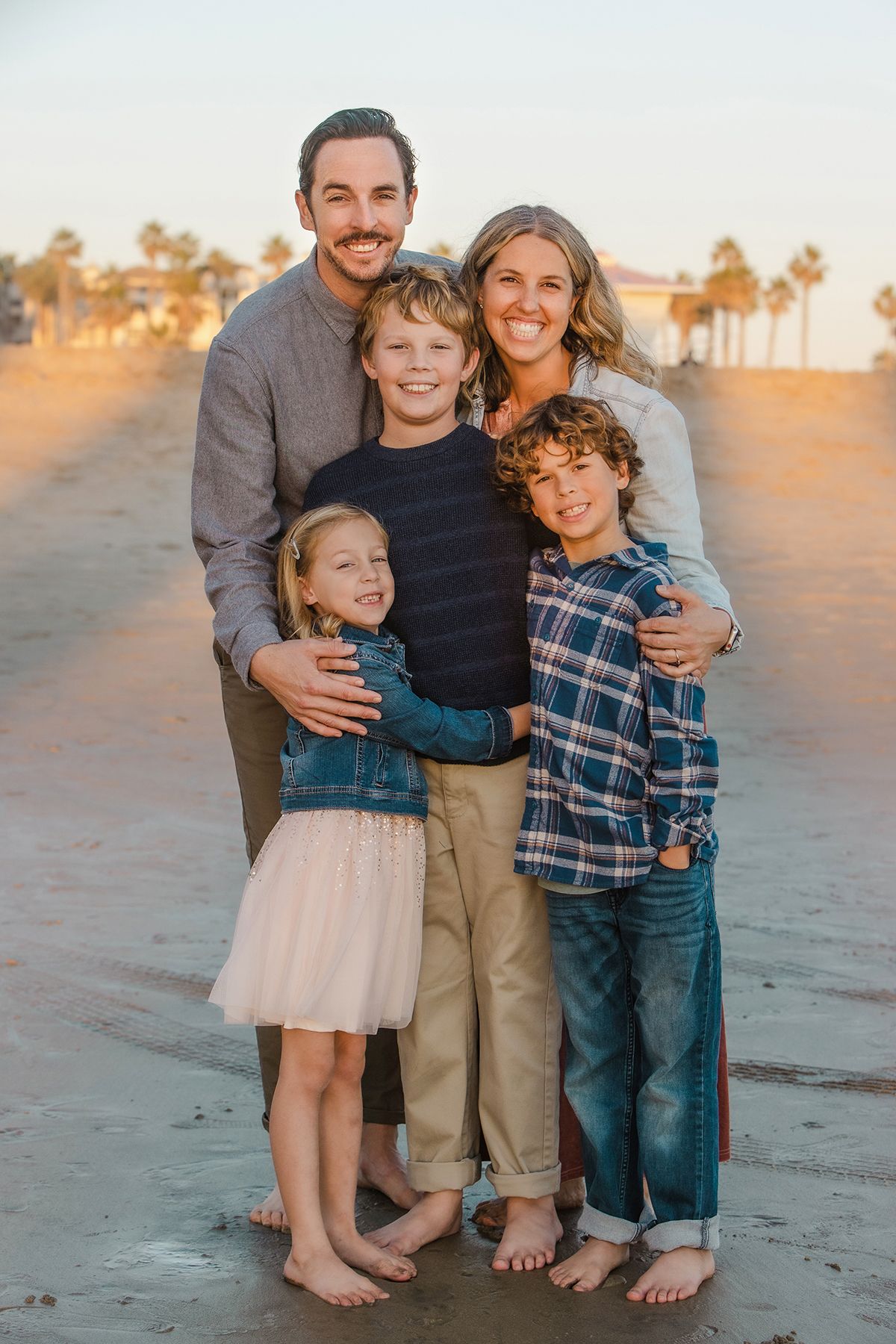 Family of five smiling together on a sandy beach at sunset with palm trees in the background.