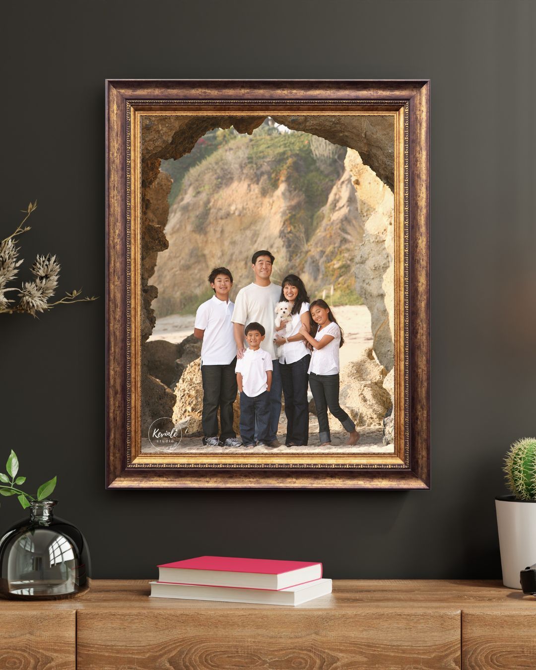 An ornate gold-framed family portrait photo displayed on a dark wall, showing parents and three children posing in a sea cave.