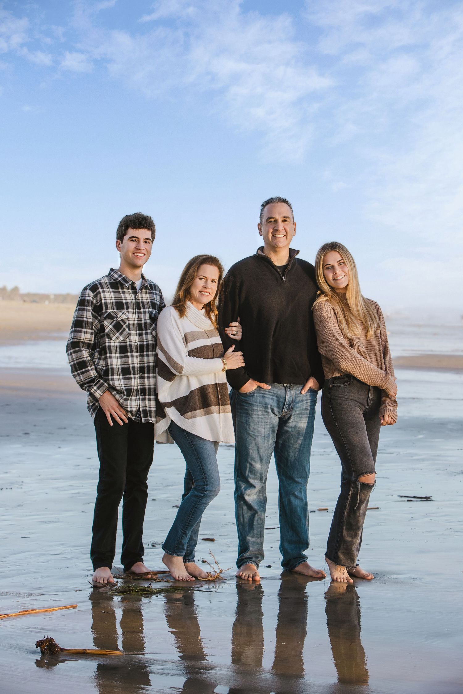 Family of four posing barefoot on a reflective beach shore under a blue sky.