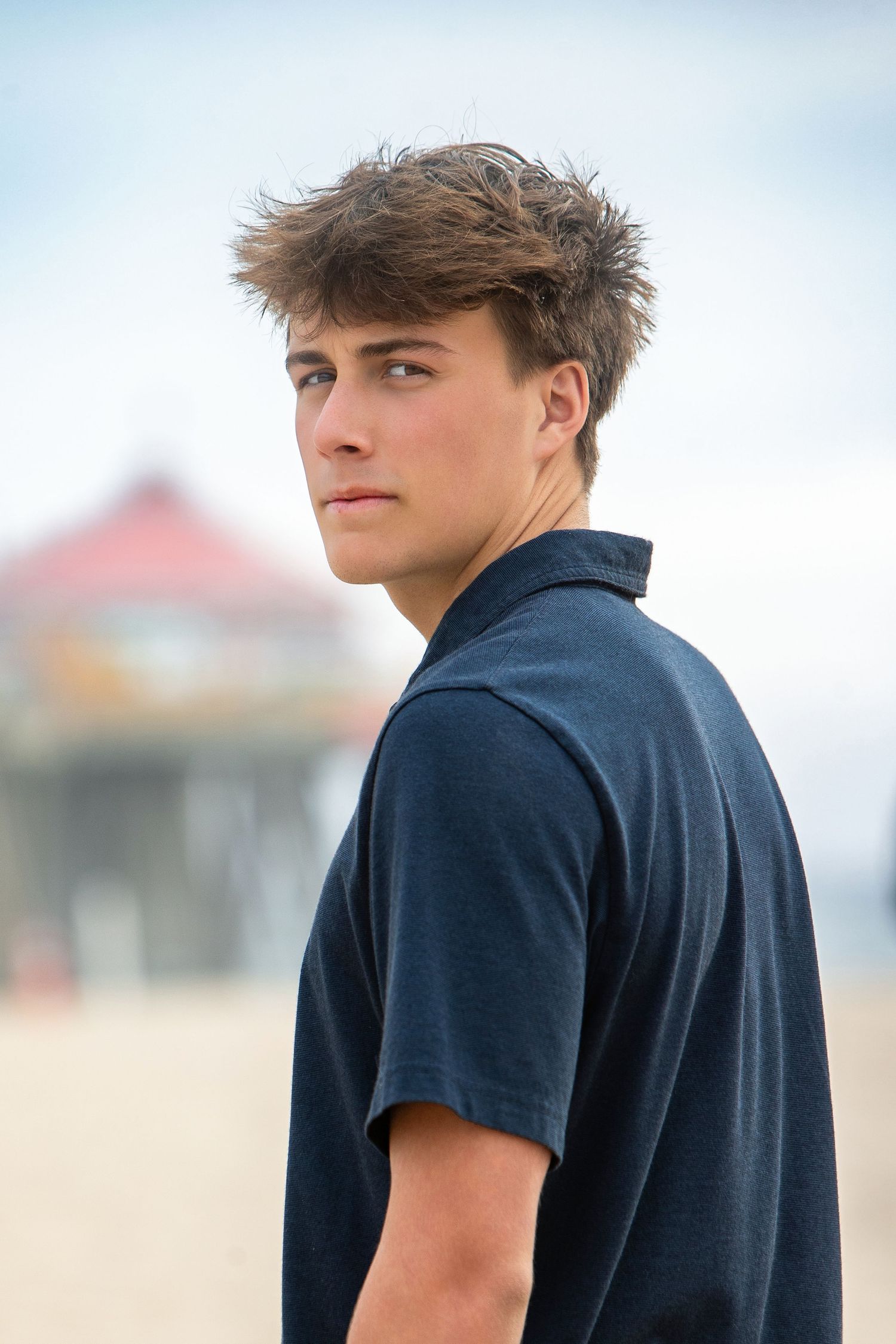 Young man in navy polo shirt posing on beach with pier in background on overcast day.