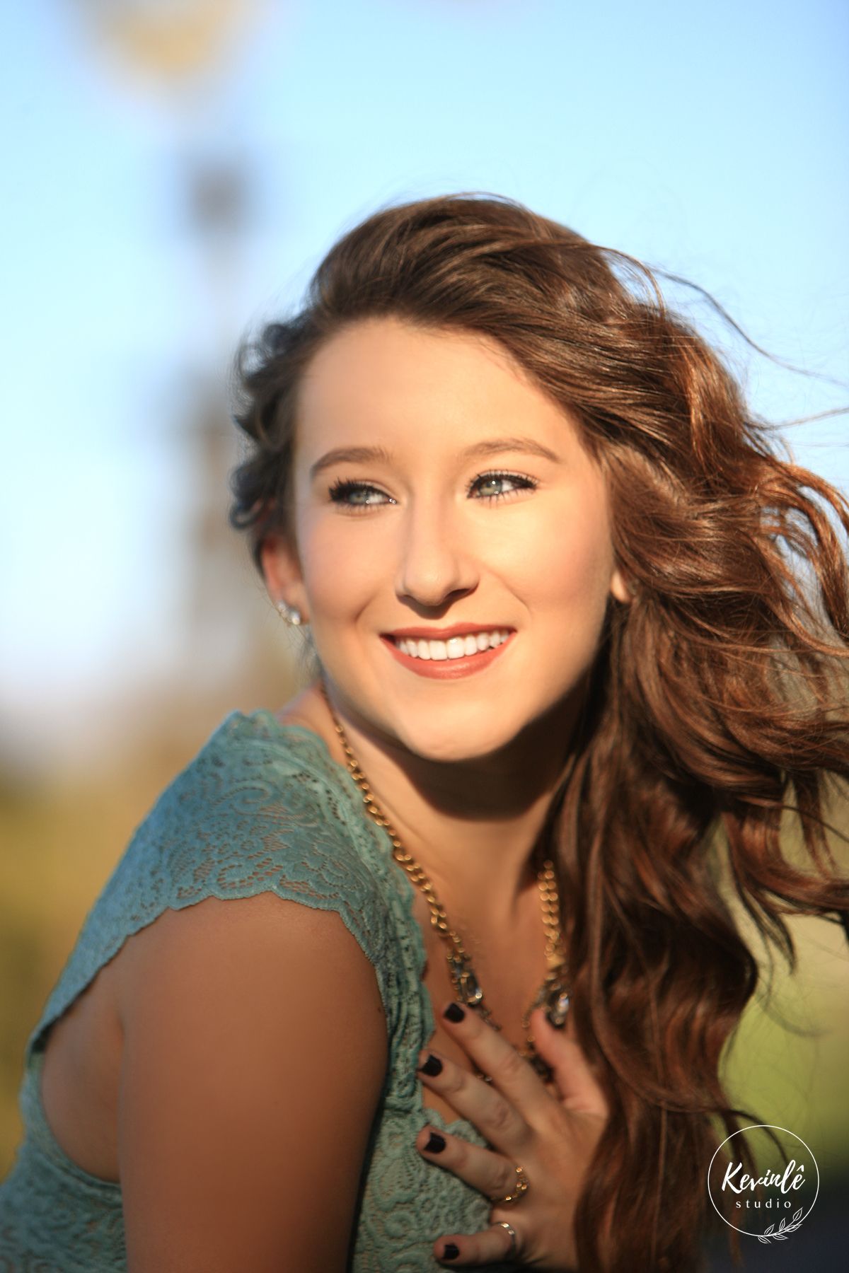 Young woman with flowing auburn hair smiles outdoors in golden light, wearing a teal lace top and gold necklace.