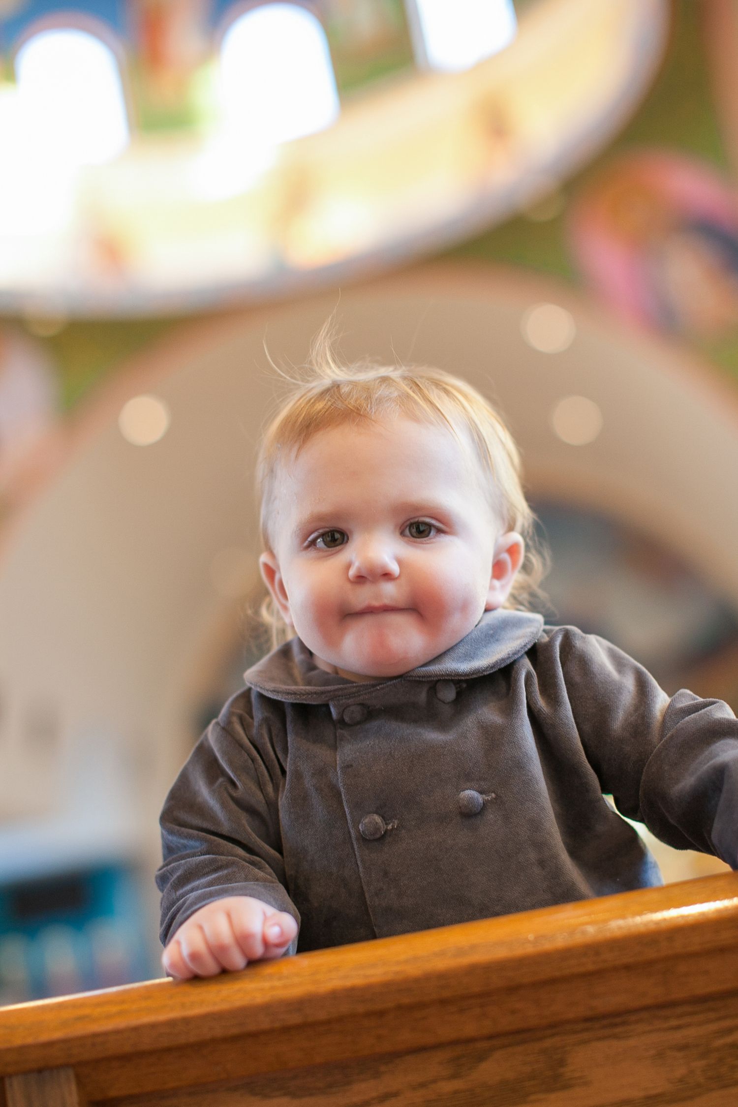 Adorable baby in gray coat standing at wooden railing, looking curiously at camera in bright indoor setting.