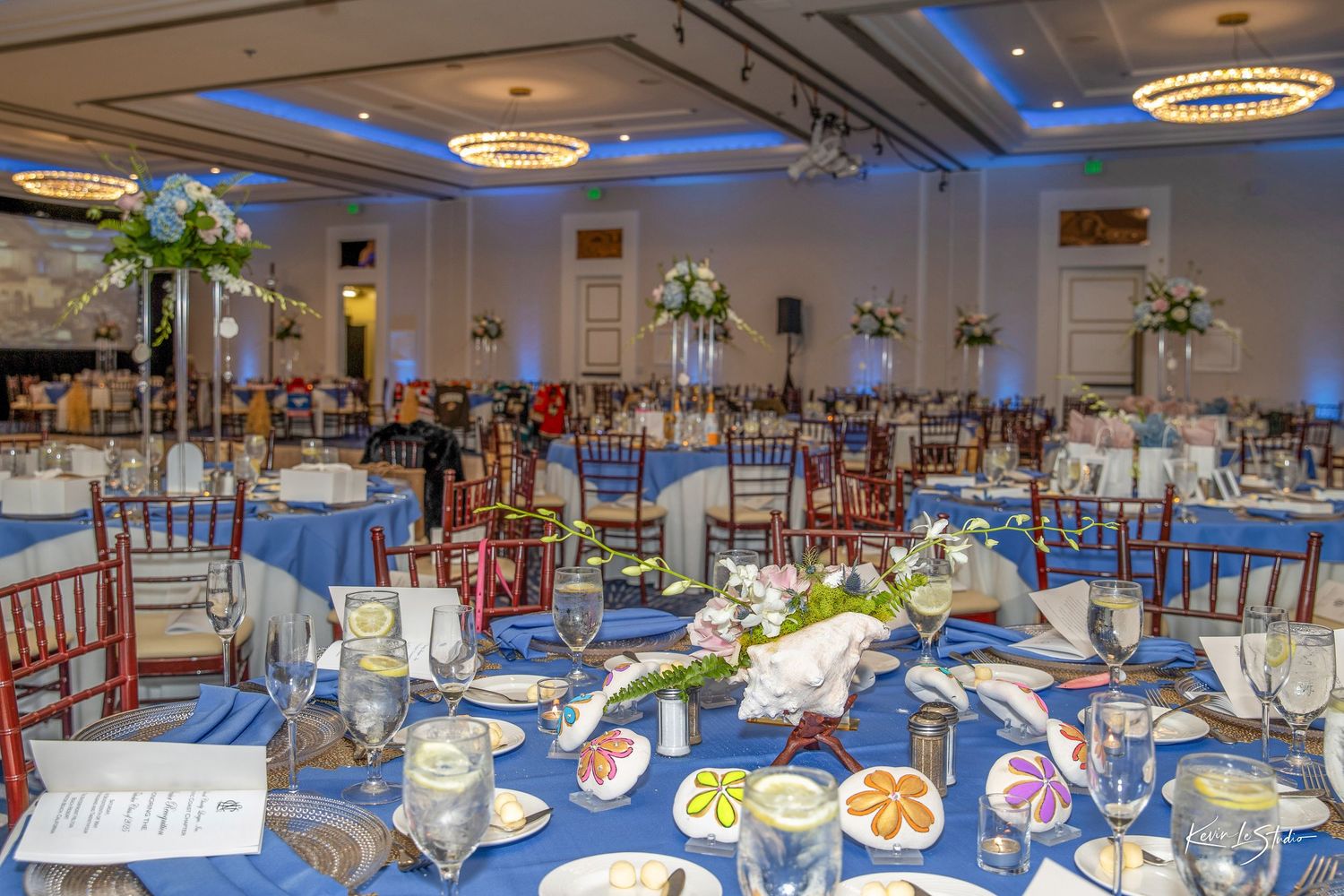 Elegant ballroom event setup with blue tablecloths, floral centerpieces, and chiavari chairs under warm chandelier lighting.