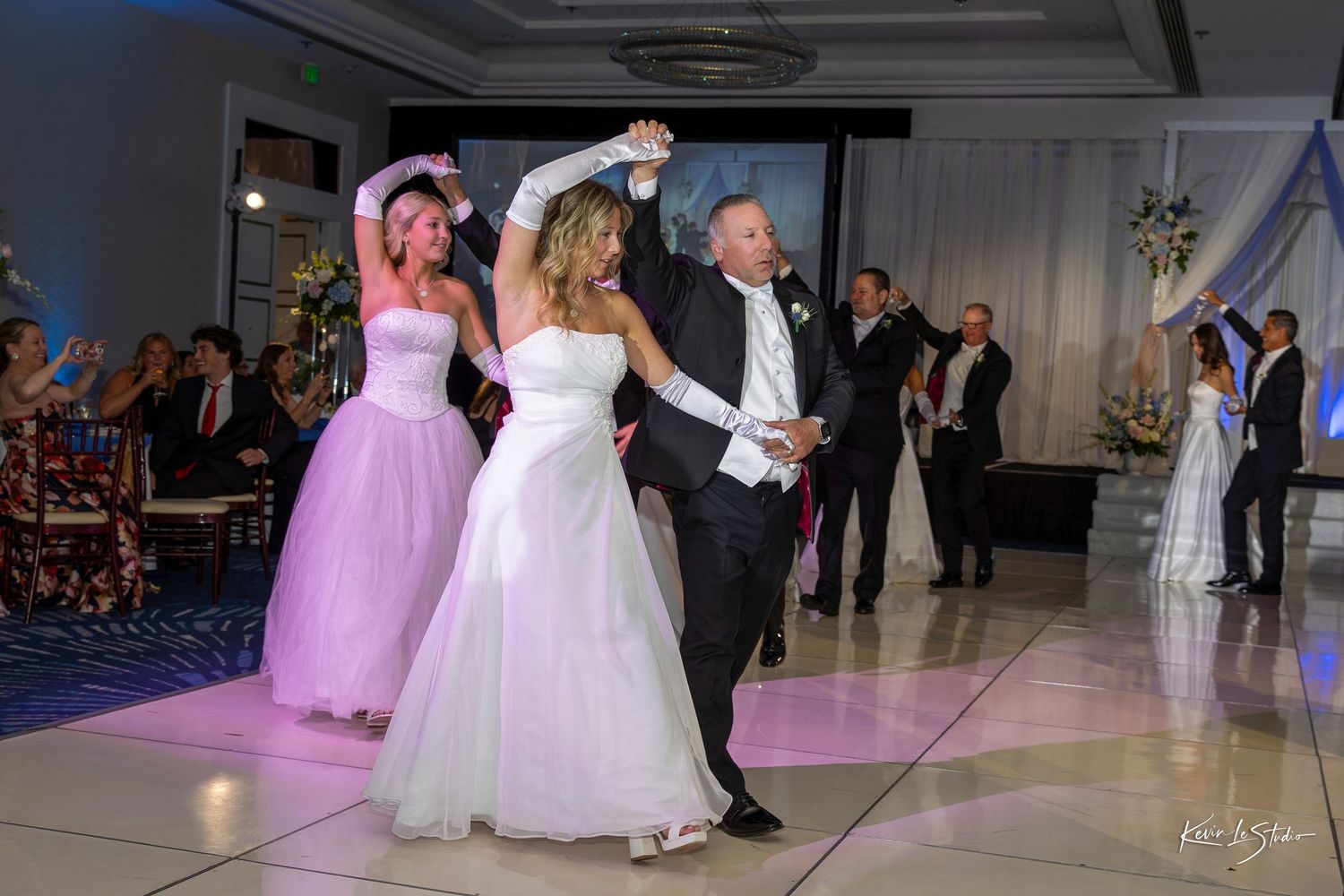 Bride and groom dancing with guests at elegant wedding reception on white dance floor.
