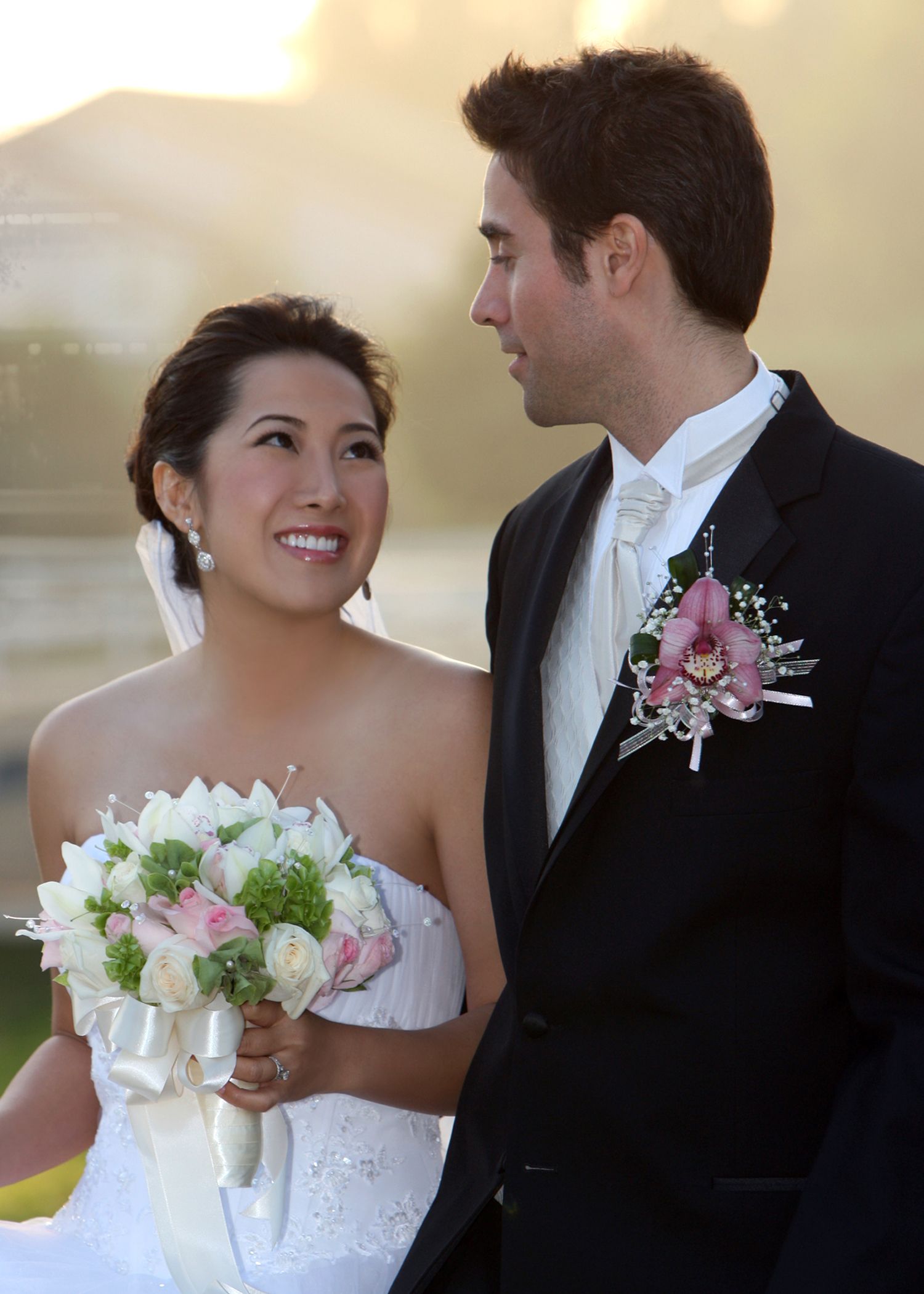 A smiling bride in a white strapless gown holds a bouquet while gazing at her groom in a black tuxedo with boutonniere.