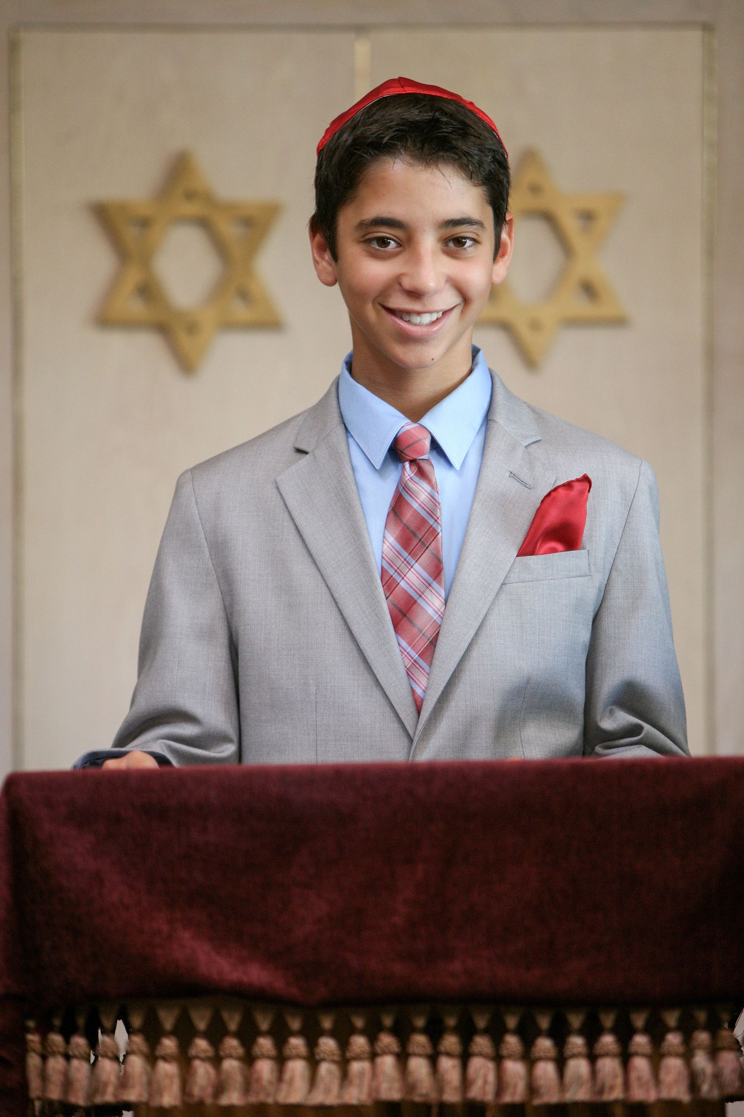 Smiling teenage boy in gray suit with red kippah stands at synagogue podium during Bar Mitzvah ceremony.