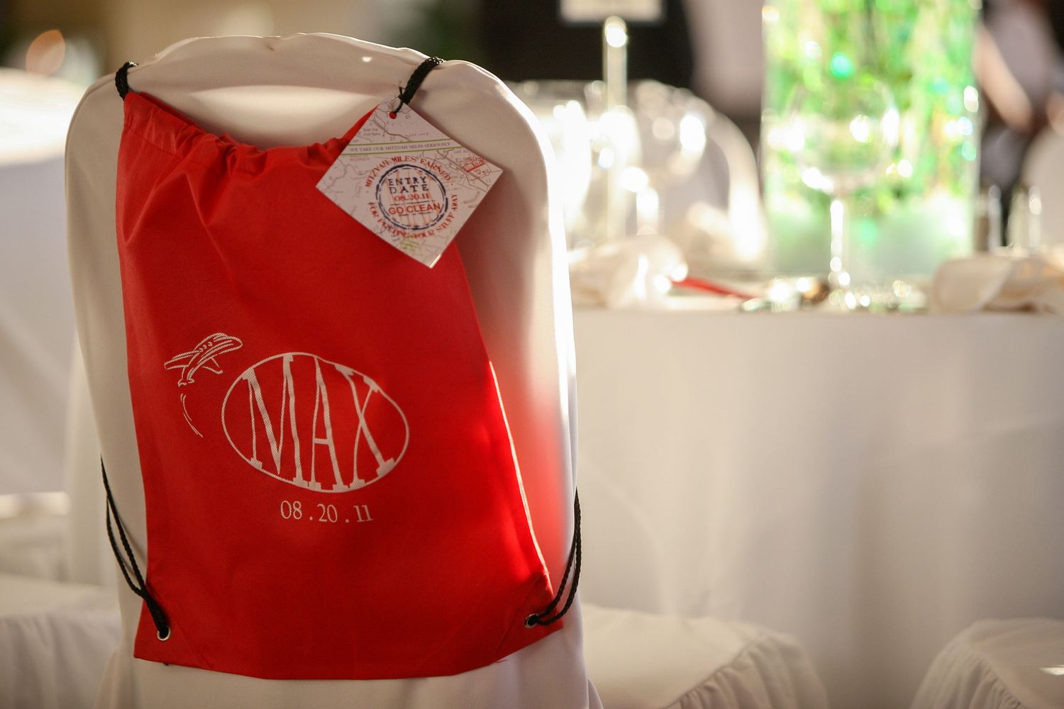 Red personalized drawstring bag with MAX monogram dated 08.20.11 hanging on white chair at elegant event.
